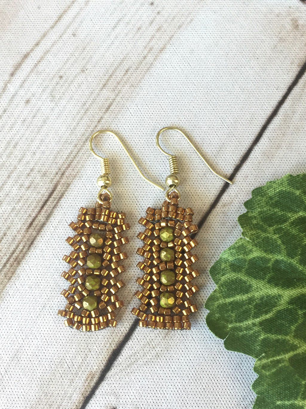 Gold seed bead bar earrings with olive crystals in the center. Laying on whitewash wood surface with a leaf to the side