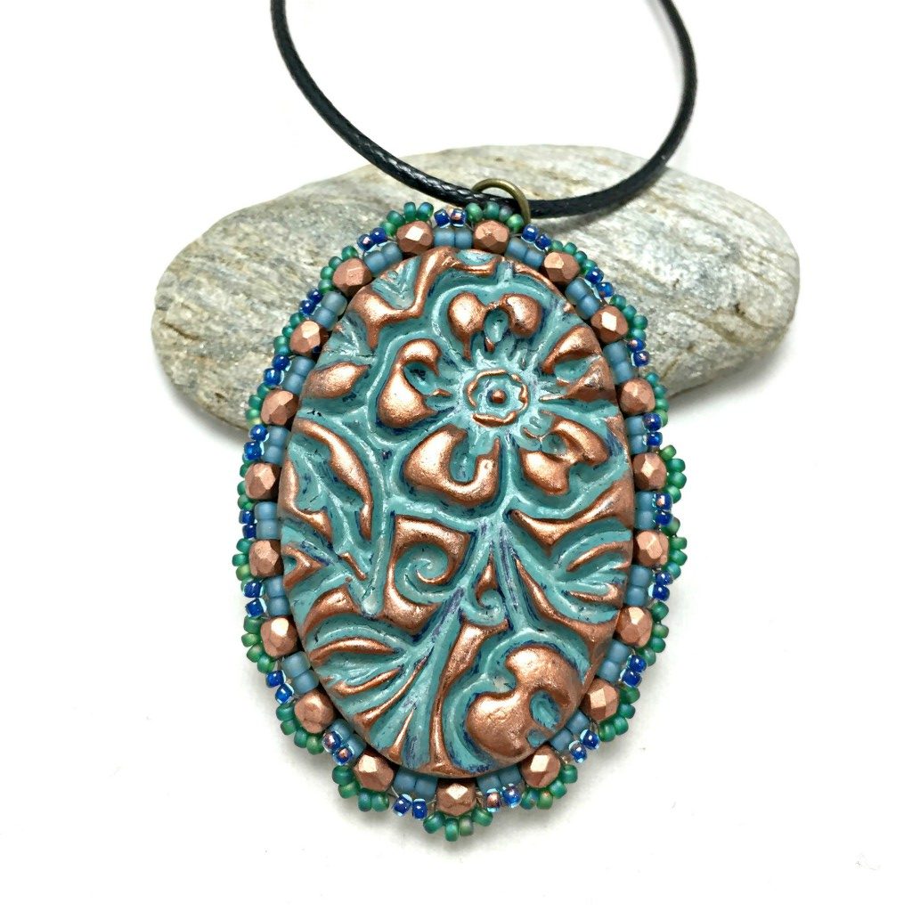 Oval floral textured pendant in patina finish with a green seed bead and copper crystal scalloped edging on a black cord necklace resting on a pebble.