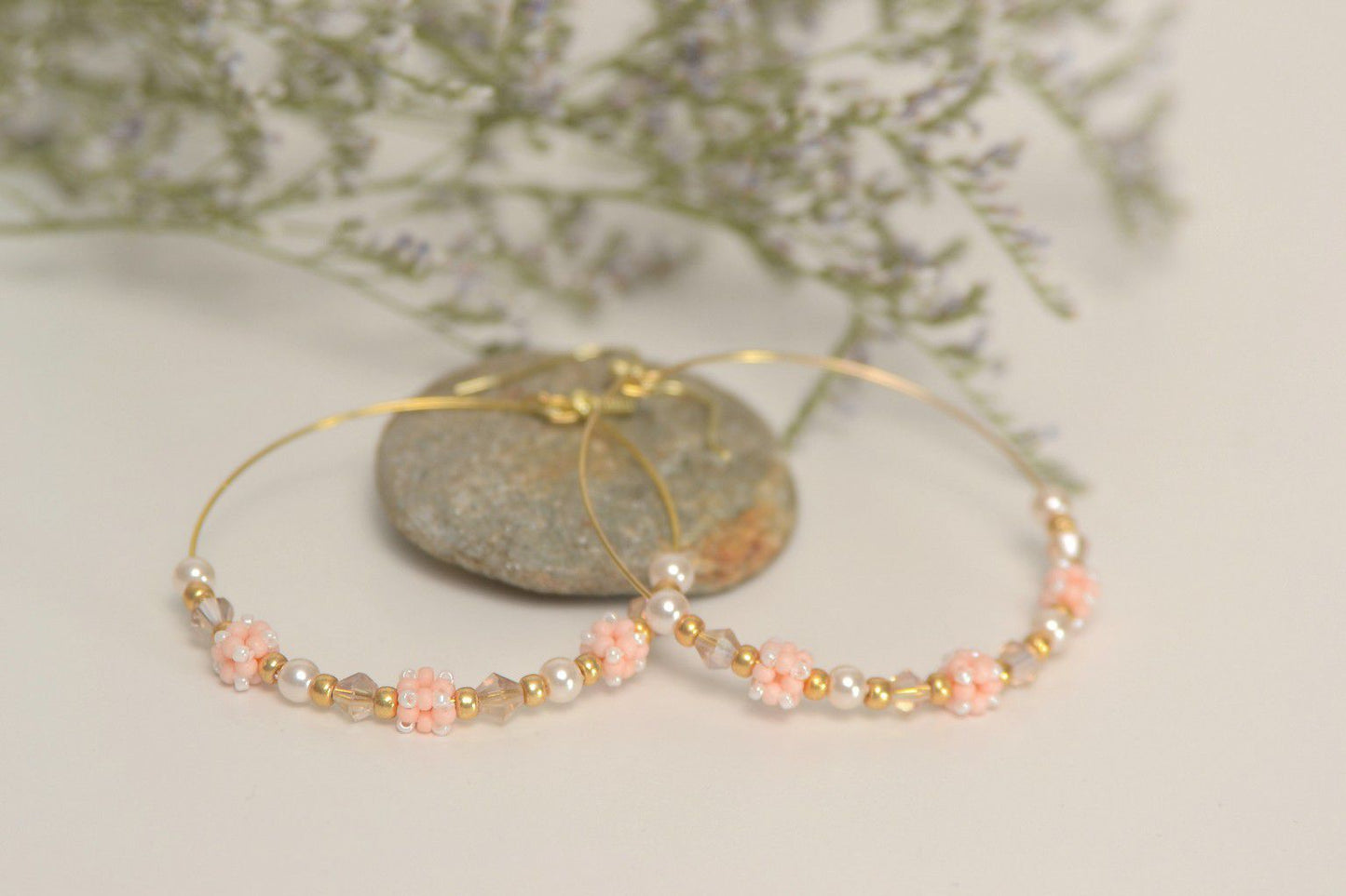 2 inch gold wire hoop earrings with peach cube beads, crystals and pearls resting on a pebble with flowers in the background.