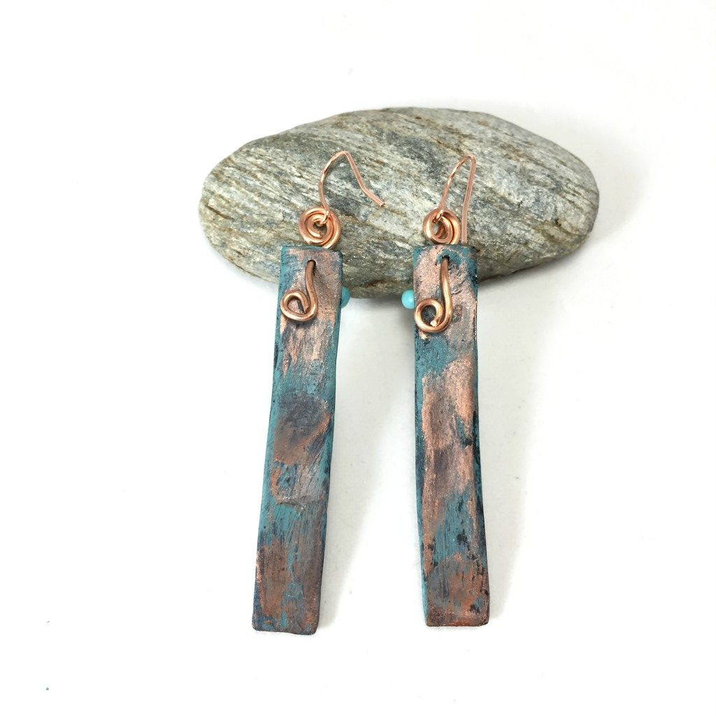 Back of long bar earrings with textured patina pattern and turquoise beads resting on a pebble.