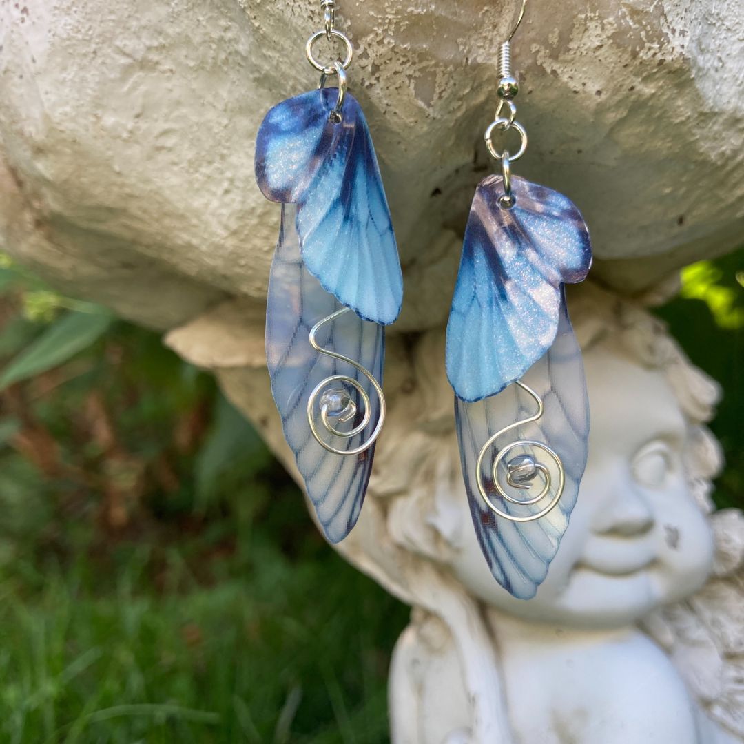 Blue butterfly double wing earrings with silver wired swirls with a silver crystal in center. On silver colored fishhook earrings photoraphed outside with a fairy garden ornament in the background.