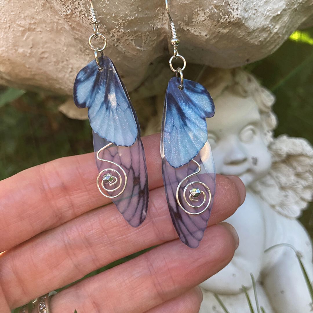Blue butterfly double wing earrings with silver wired swirls with a silver crystal in center. On silver colored fishhook earrings photoraphed  held in a hand with a fairy garden ornament in the background.
