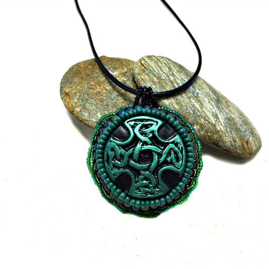 Green and black Celtic cross pendant with green twisted beaded edging resting on a pebble.