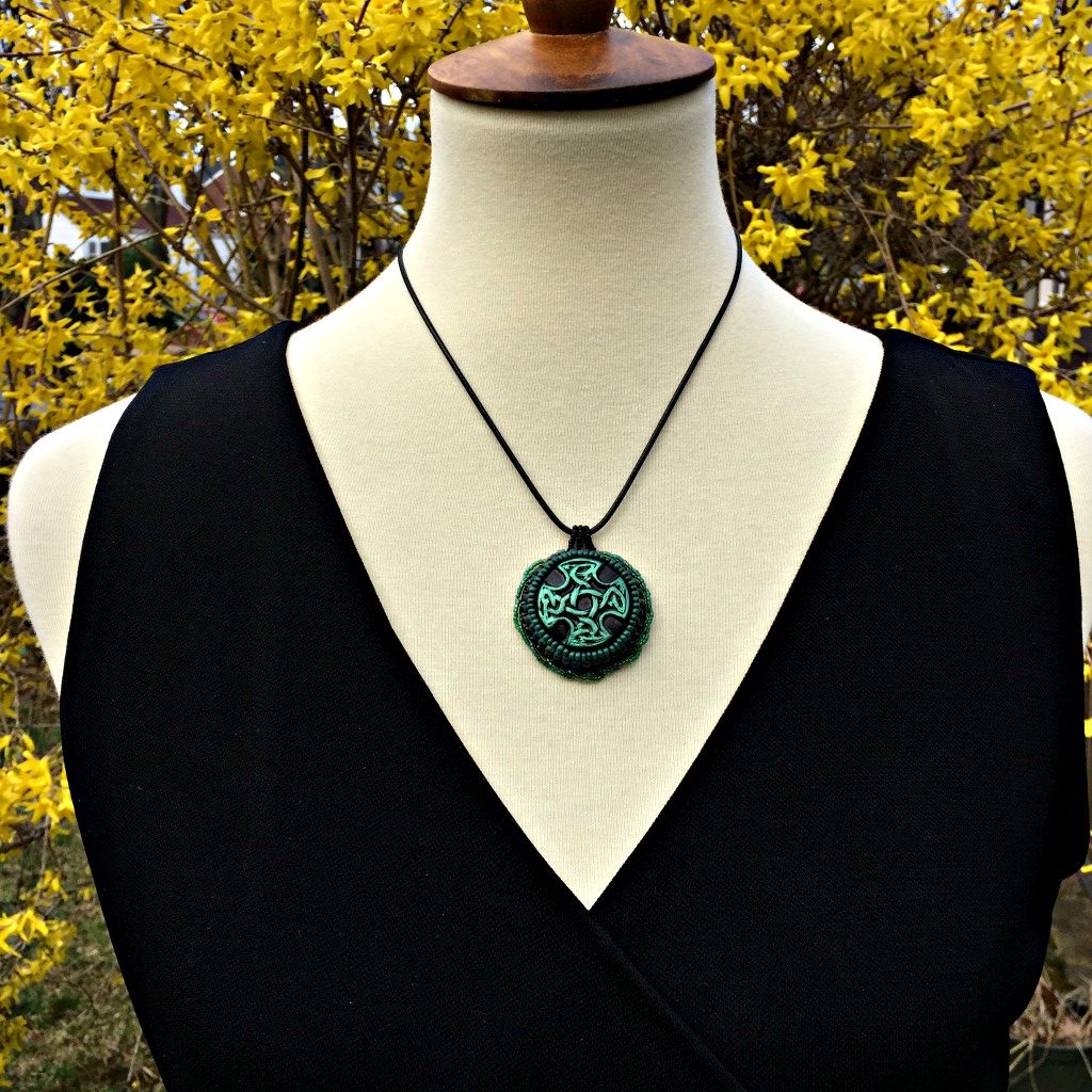 Green and black Celtic cross pendant with green twisted beaded edging displayed on a model outside in a garden with a forsythia tree in the background.