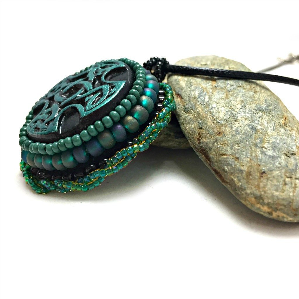 Side profile of green and black Celtic cross pendant with green twisted beaded edging resting on a pebble.