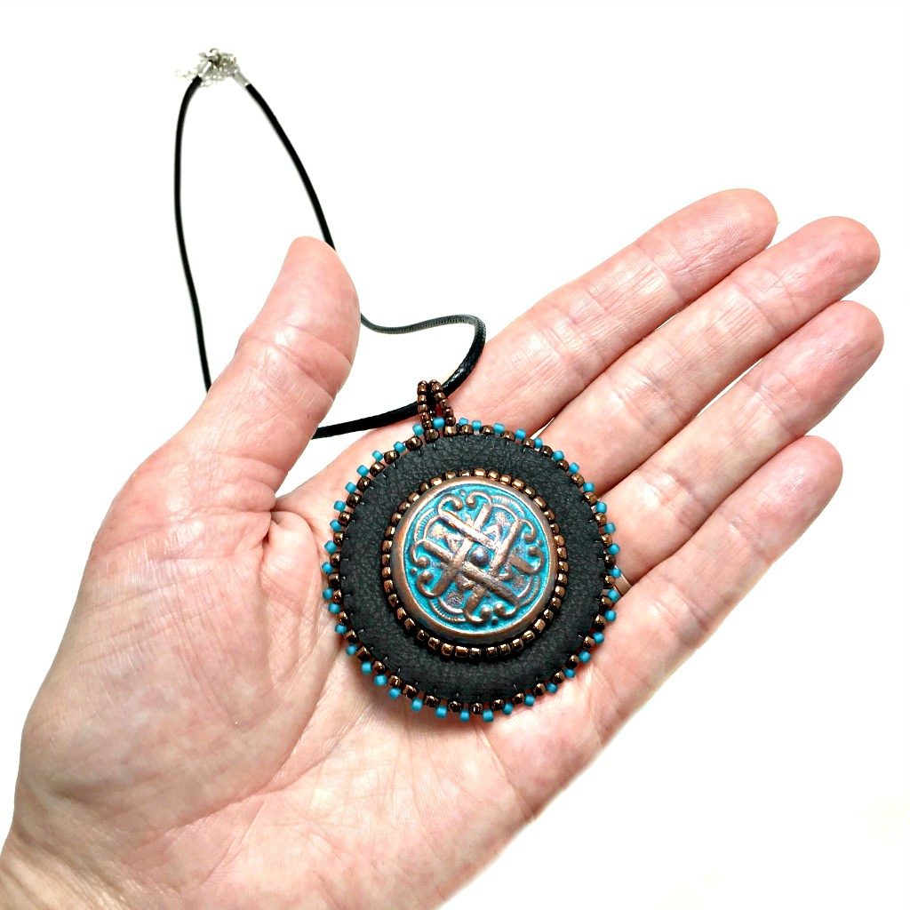Black faux leather round pendant with beaded edge in gold and turquoise with a patina Celtic knot motif in the center with beading. Necklace is on a black cord chain held in a hand for size reference.