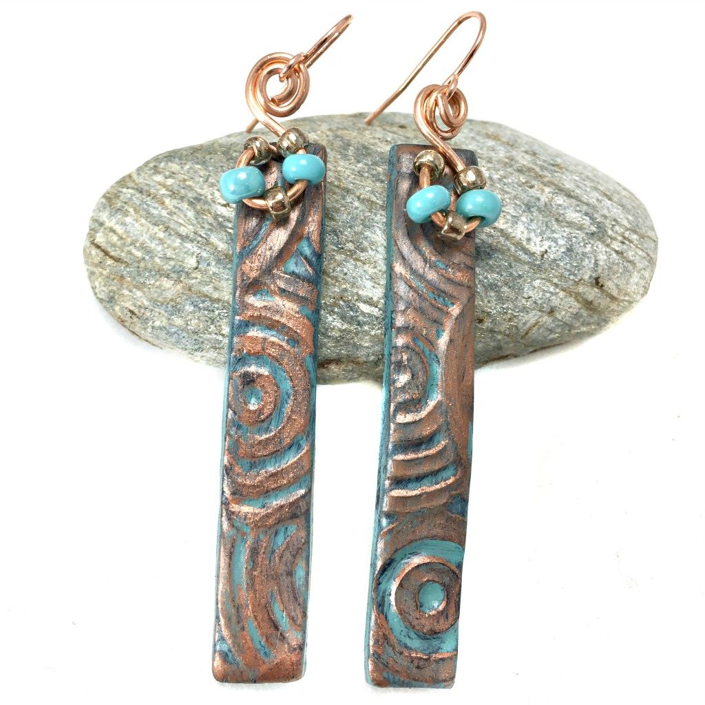 Long bar earrings with textured patina pattern and turquoise beads resting on a pebble.