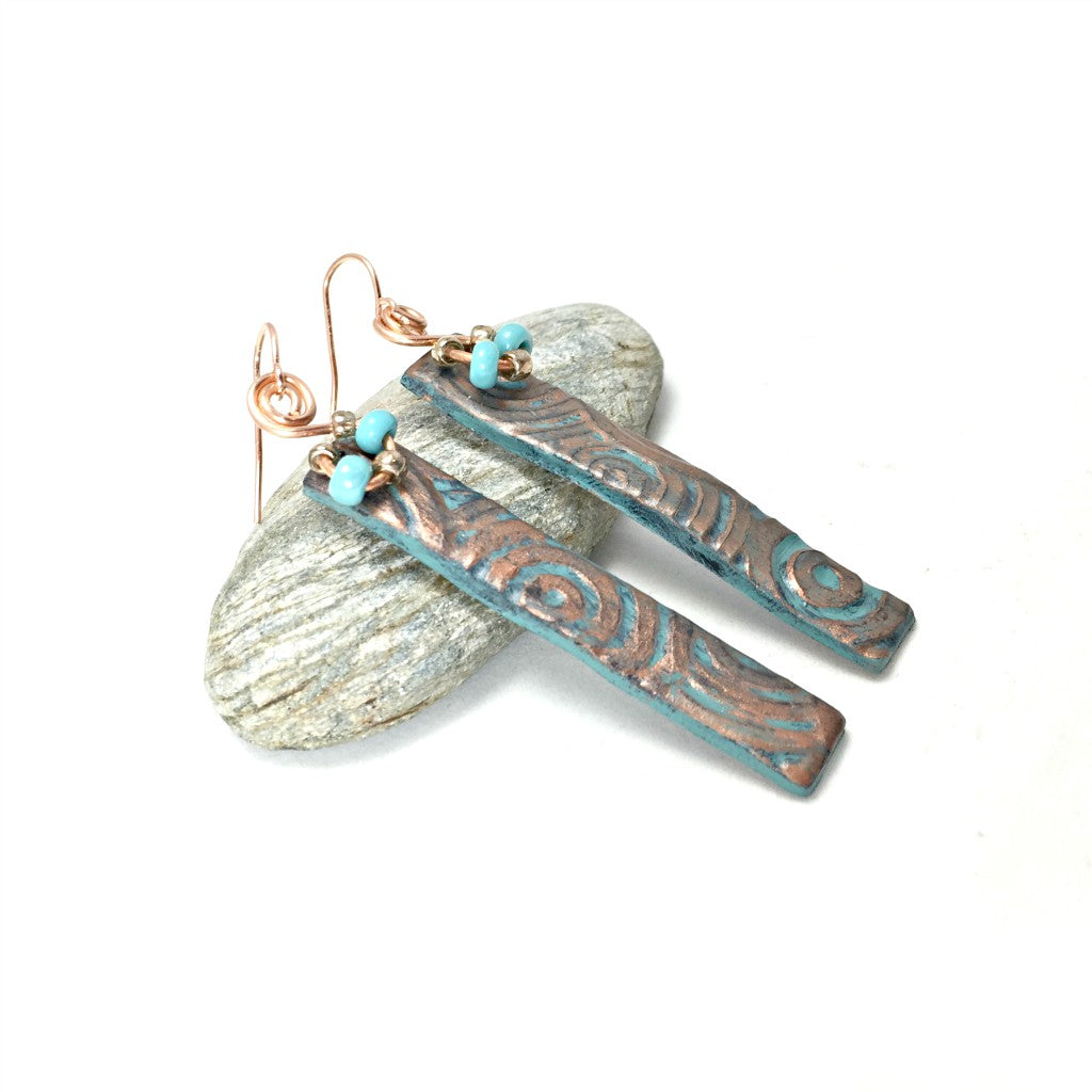 Side view of long bar earrings with textured patina pattern and turquoise beads resting on a pebble.