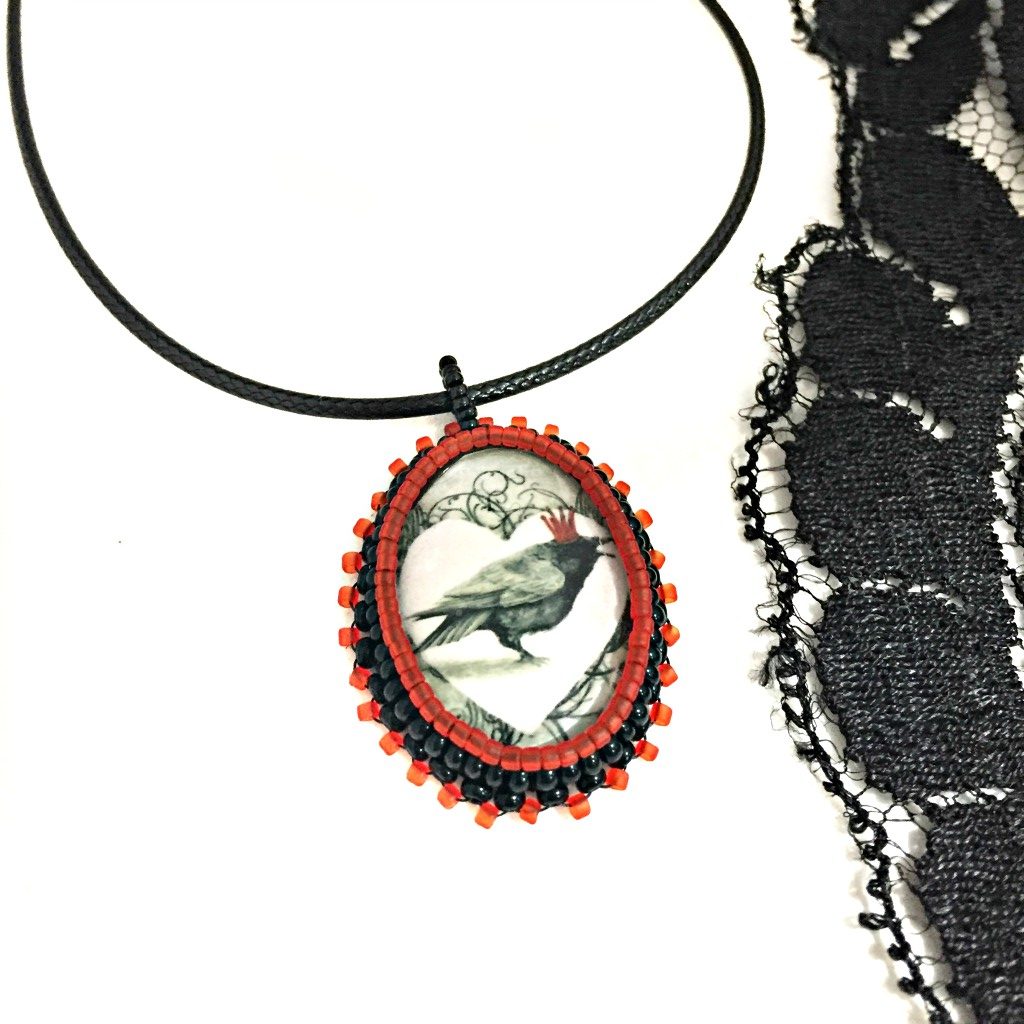 Oval red and black beaded pendant necklace with graphic of a raven wearing a red crown inside a white heart with a piece of black lace to the right hand side, the photo