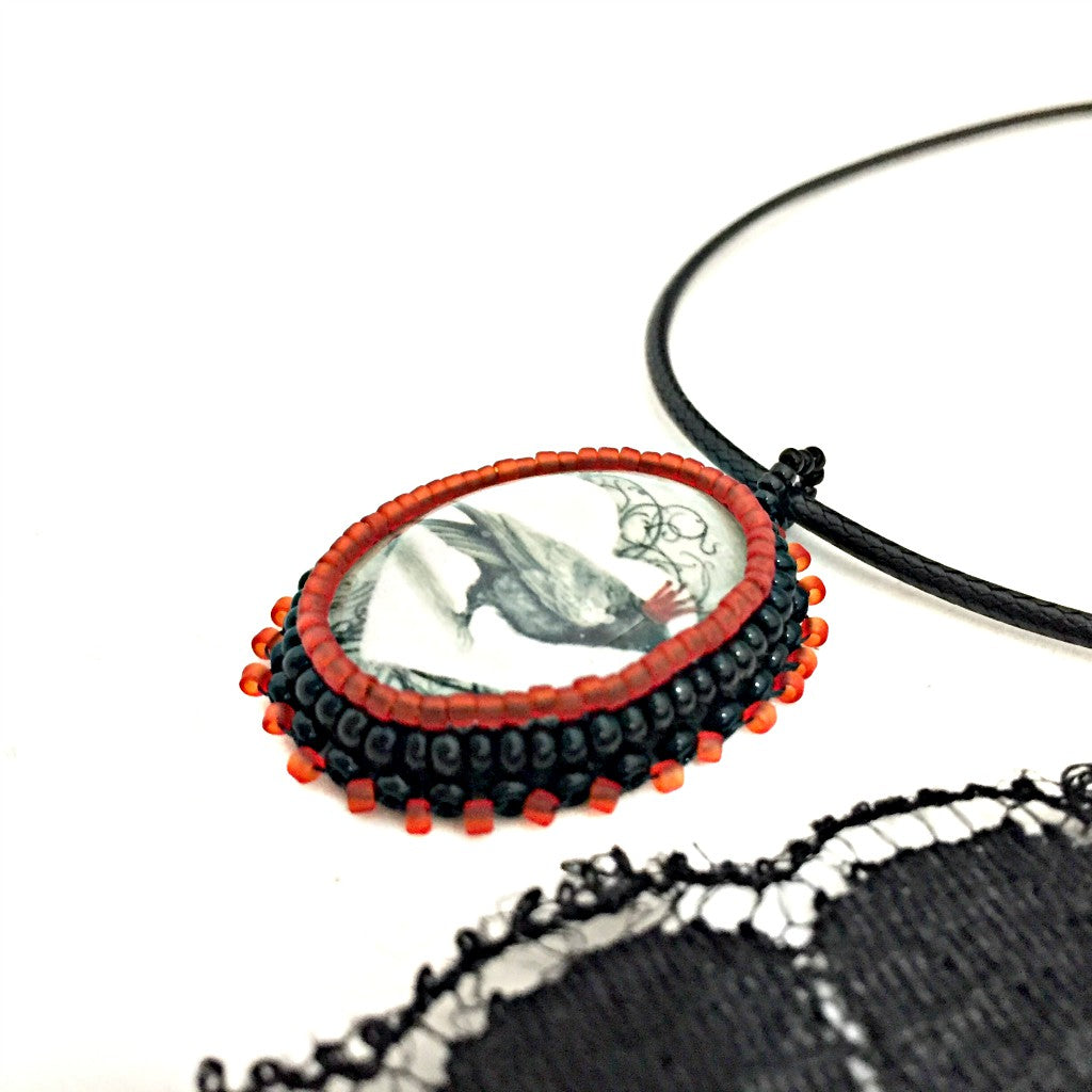 Side view of an oval red and black beaded pendant
necklace with graphic of a raven wearing a red crown inside a white heart. A piece of black lace is to the right hand side.