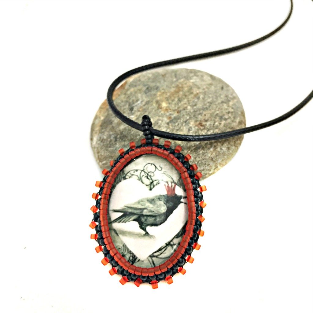 Oval red and black beaded pendant necklace with graphic of a raven wearing a red crown inside a white heart resting on a pebble.