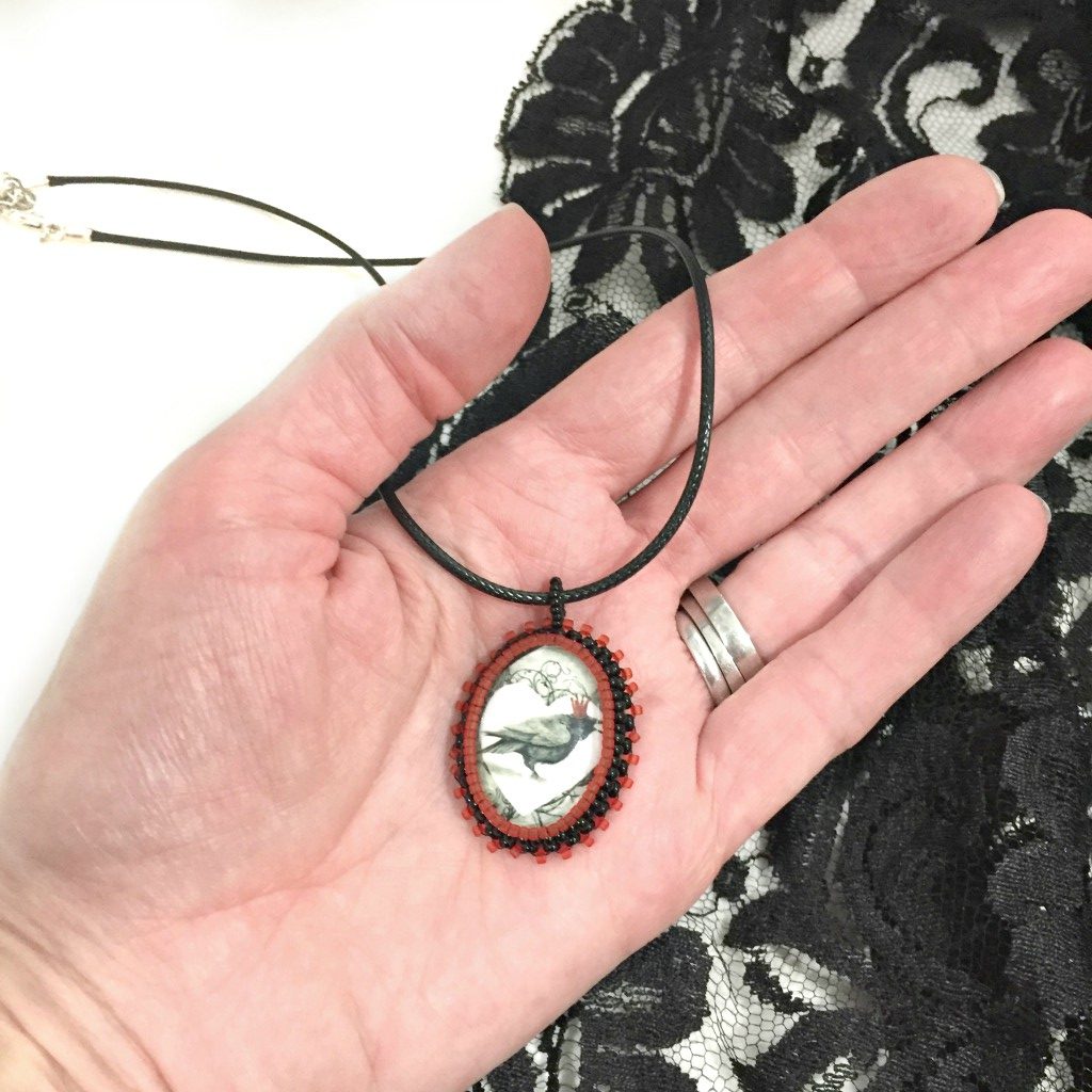 Oval red and black beaded pendant necklace with graphic of a raven wearing a red crown inside a white heart held in a hand with black lace in the background.