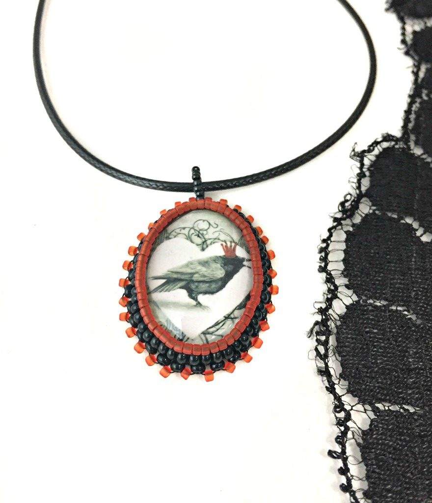 Oval red and black beaded pendant necklace with graphic of a raven wearing a red crown inside a white heart with a piece of black lace to the right hand side, the photo