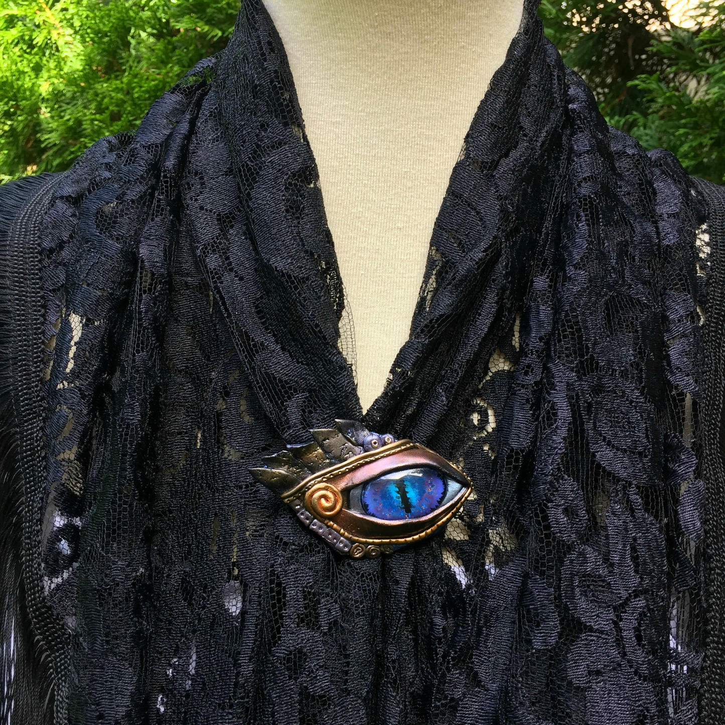 A polymer clay egyptian blue eye brooch with gold, silver feather and swirl details on a lace shawl on model.