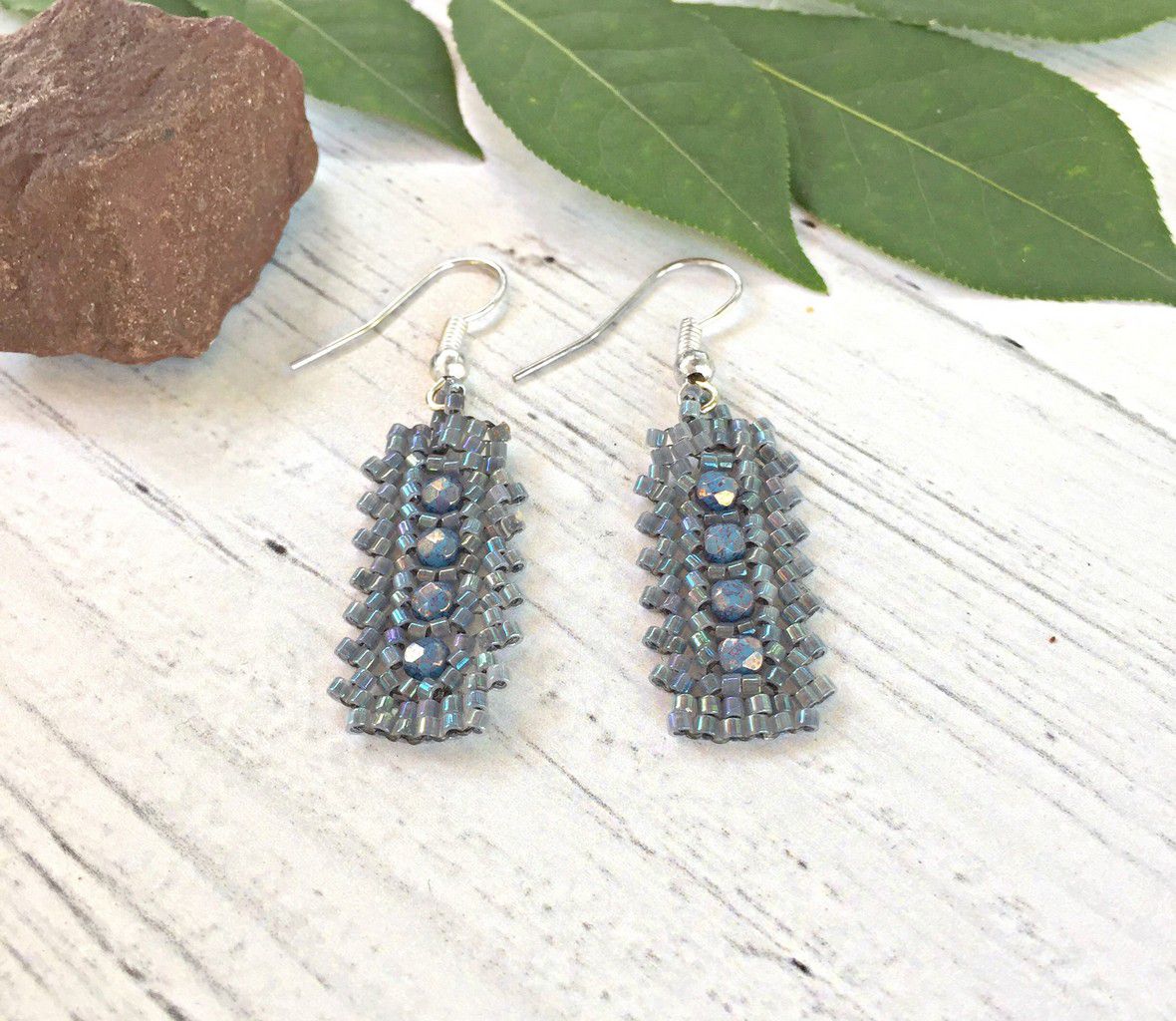 Gray seed bead lace rectangle earrings. With blue crystals in the center laying flat on white was wood surface with a rock and leaf in the background