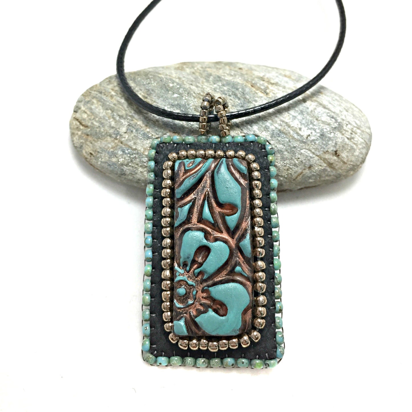 Close-up of front view of patina faux leather floral textured rectangle pendant necklace resting on a pebble.