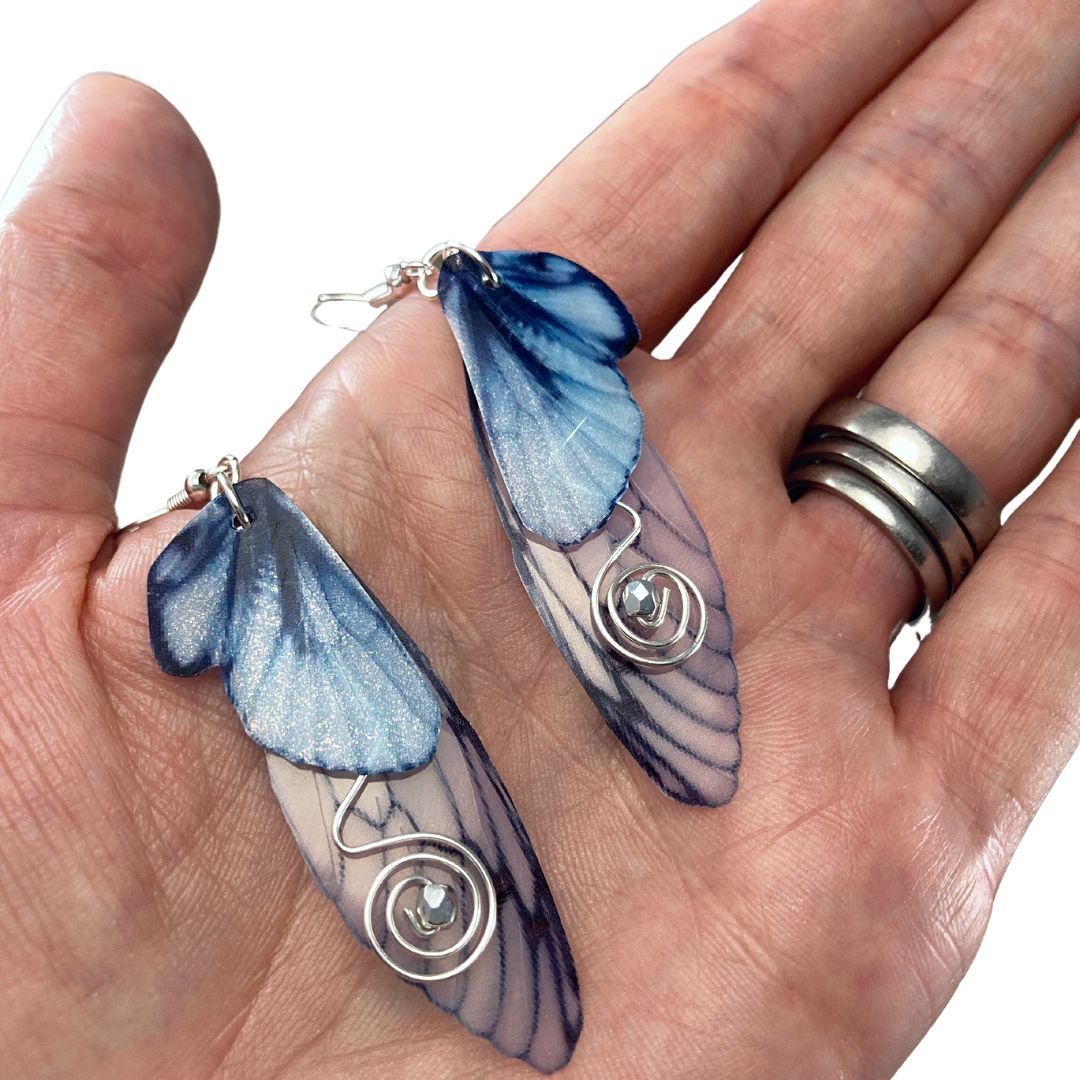 Blue butterfly double wing earrings with silver wired swirls with a silver crystal in center. On silver colored fishhook earrings photoraphed in the palm on a hand.