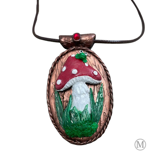 Oval pendant with red spotted mushroom with grass and moss at stem with little green frog sitting on top of the mushroom. Brown cord necklace through a tube bail with a red crystal