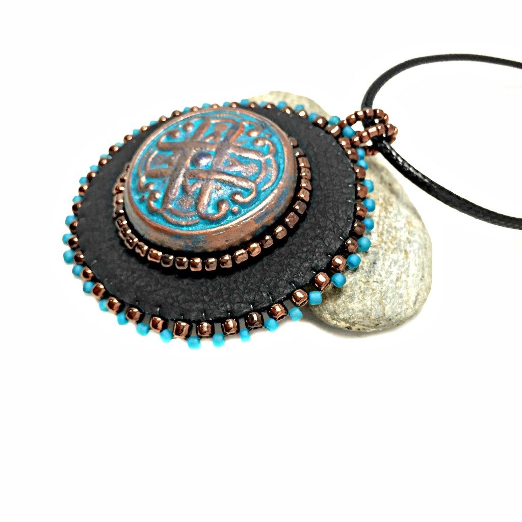 Side profile of black faux leather round pendant with beaded edge in gold and turquoise with a patina Celtic knot motif in the center with beading. Necklace is on a black cord chain and is resting on a rock.