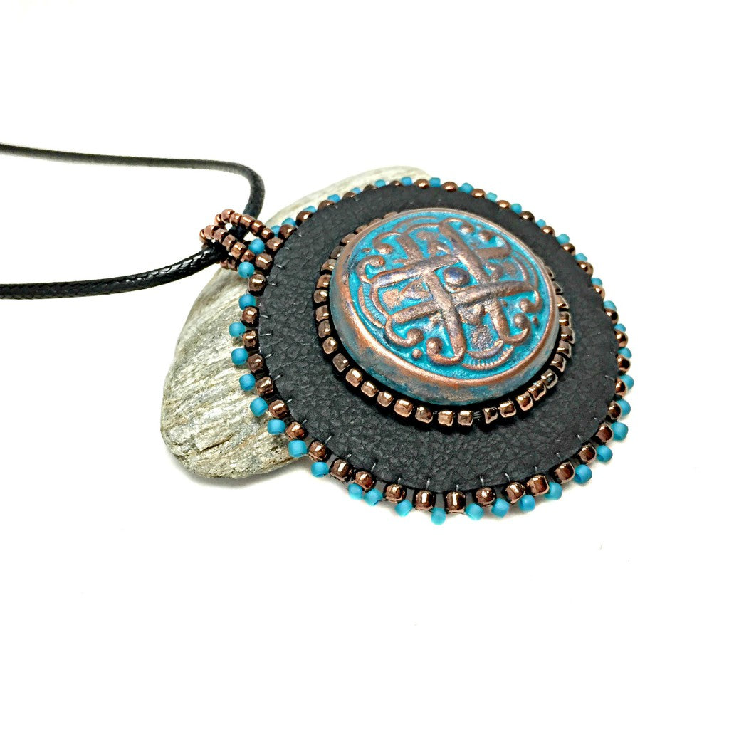 Close up of black faux leather round pendant with beaded edge in gold and turquoise with a patina Celtic knot motif in the center with beading. Necklace is on a black cord chain.