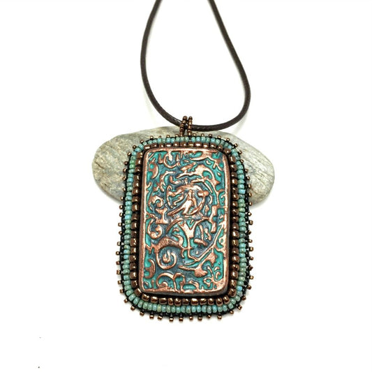 Large copper patina textured floral Rectangle pendant with turquoise and bronze beaded edging on a brown cord necklace chain.