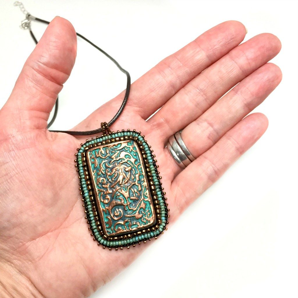 Large copper patina textured floral Rectangle pendant with turquoise and bronze beaded edging on a brown cord necklace chain held in a hand for size reference.