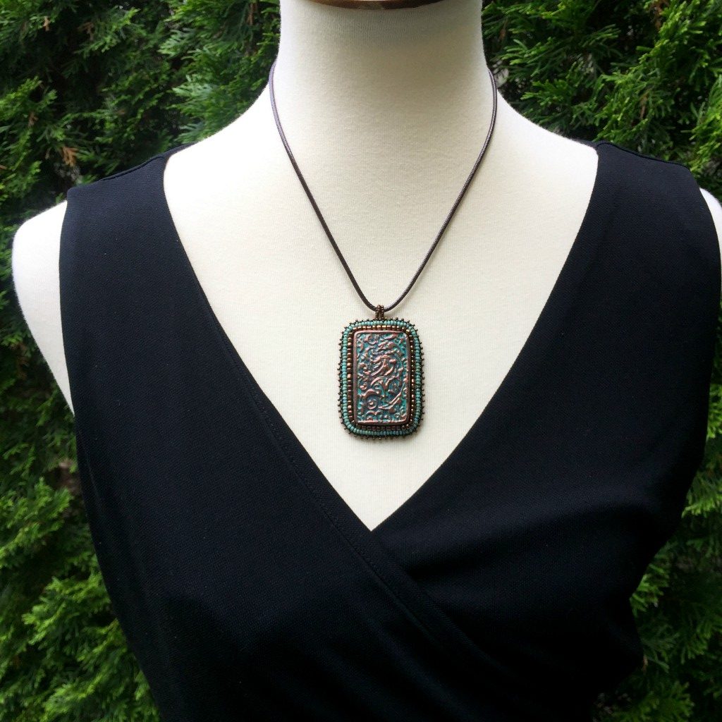 Model wearing large copper patina textured floral Rectangle pendant with turquoise and bronze beaded edging on a brown cord necklace chain.