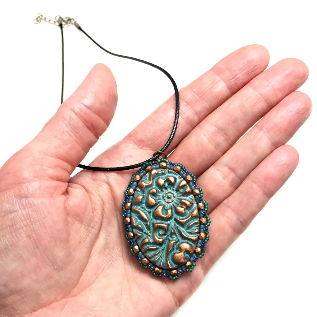 Oval floral textured pendant in patina finish with a green seed bead and copper crystal scalloped edging on a black cord necklace resting on a pebble held in a hand for size reference.