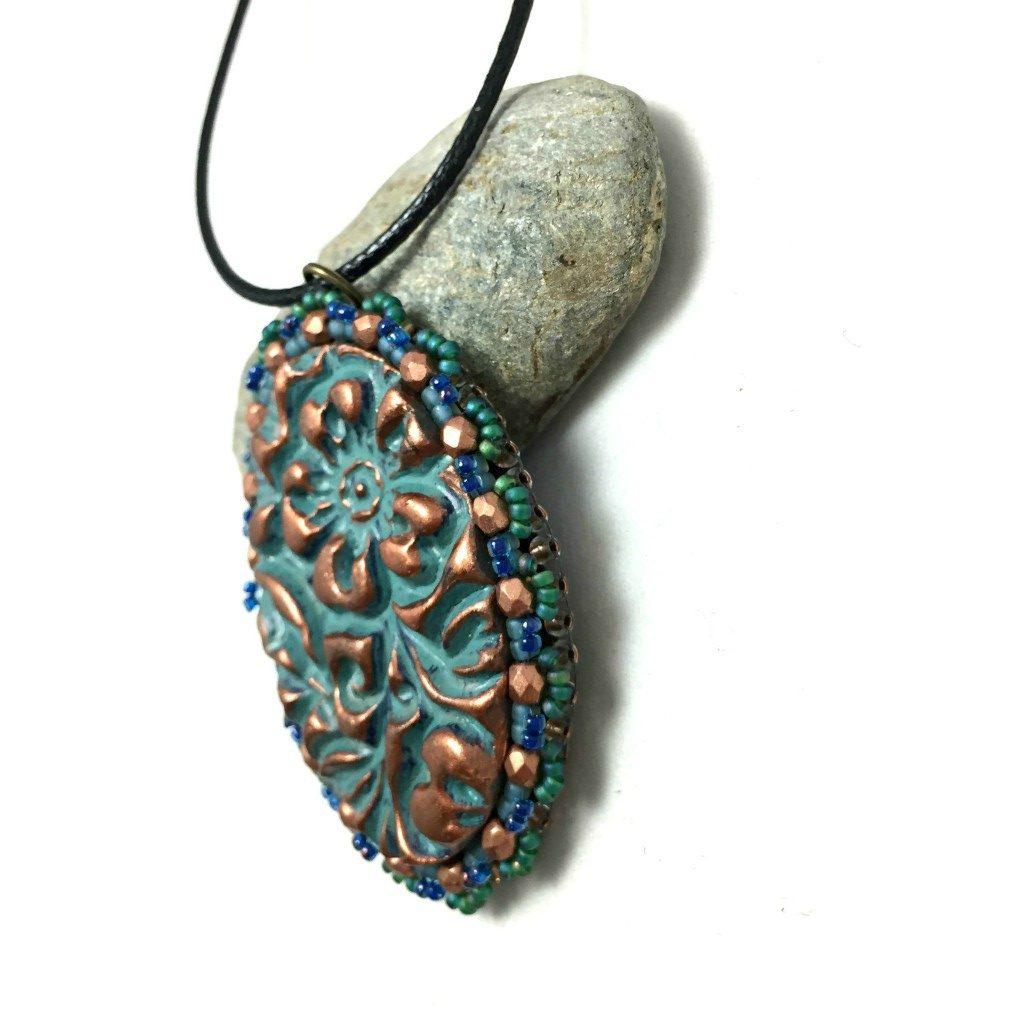 Left side profile of oval floral textured pendant in patina finish with a green seed bead and copper crystal scalloped edging on a black cord necklace resting on a pebble.
