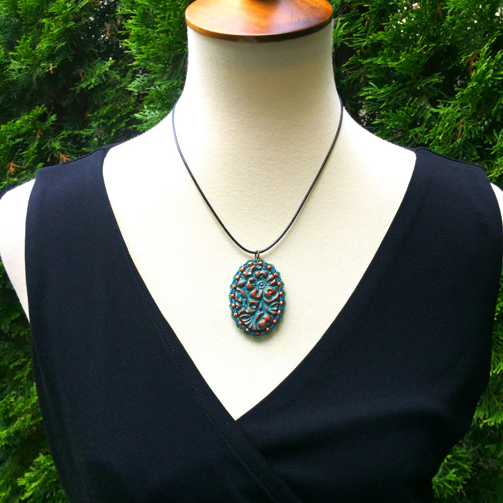 A model wearing an oval floral textured pendant in patina finish with a green seed bead and copper crystal scalloped edging on a black cord necklace resting on a pebble.