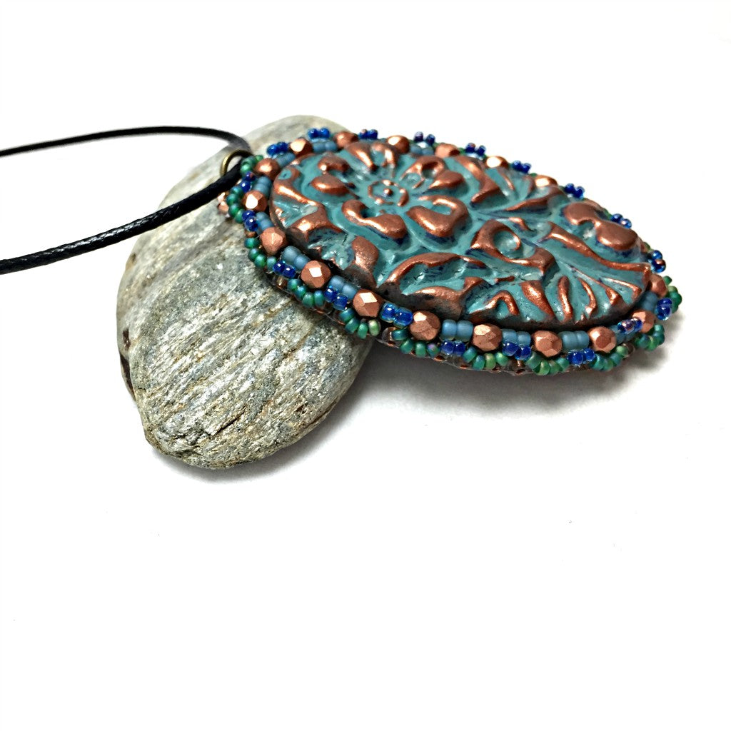 Right side profile of oval floral textured pendant in patina finish with a green seed bead and copper crystal scalloped edging on a black cord necklace resting on a pebble.