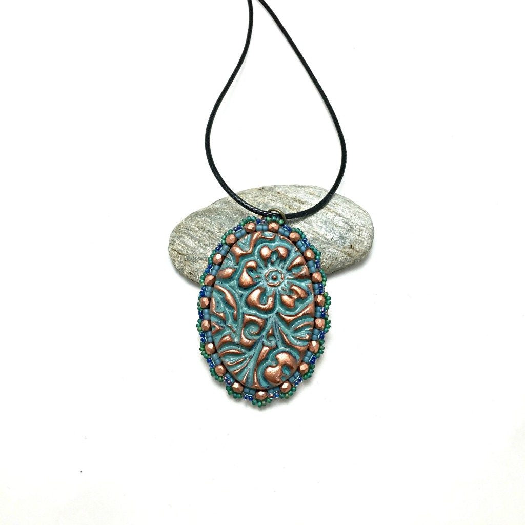 Oval floral textured pendant in patina finish with a green seed bead and copper crystal scalloped edging on a black cord necklace resting on a pebble.
