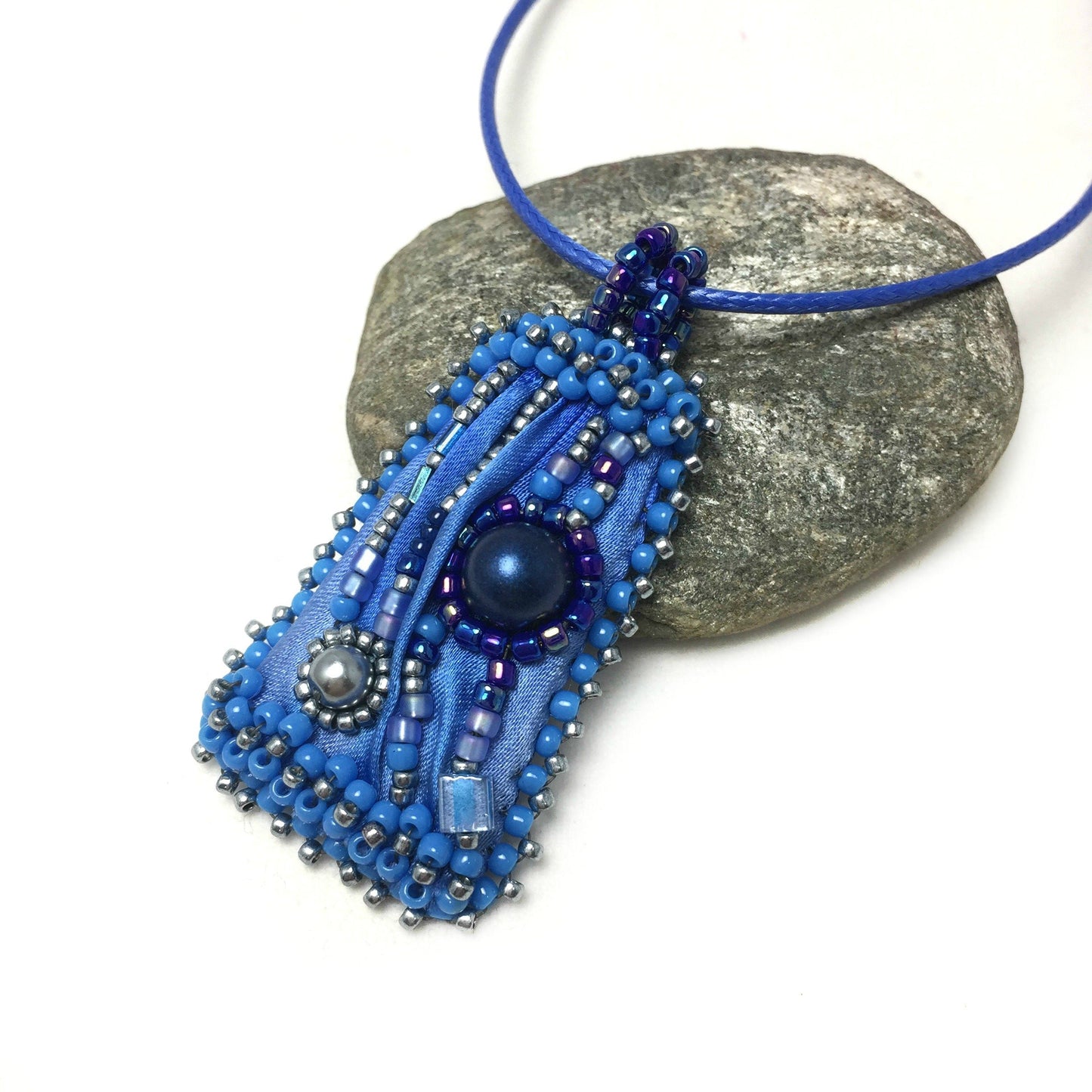 Small geometric, blue silk beaded pendant necklace resting on a pebble.