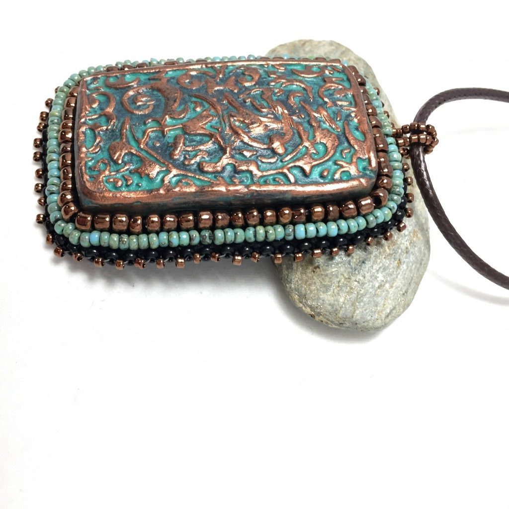 Left view of large copper patina textured floral Rectangle pendant with turquoise and bronze beaded edging on a brown cord necklace chain.