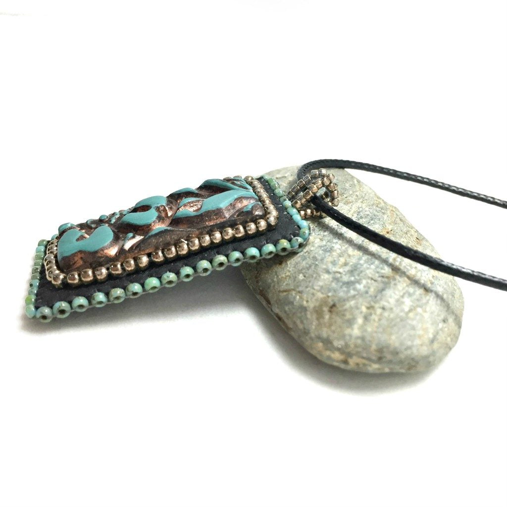 side view of faux leather patina rectangle floral beaded pendant resting on a pebble.