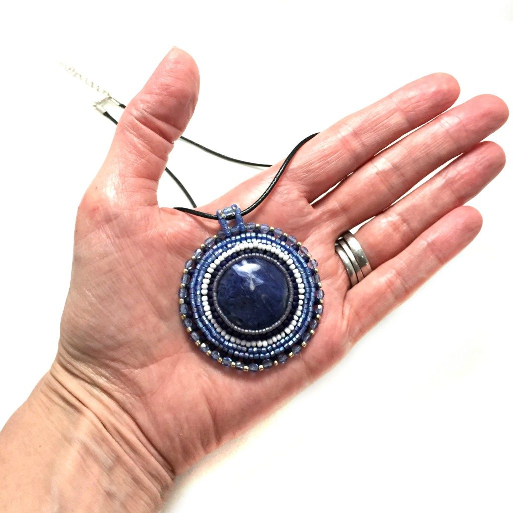 close up of round blue sodalite gemstone necklace with blue, silver and white seed bead edging. necklace is held in a hand for size reference.