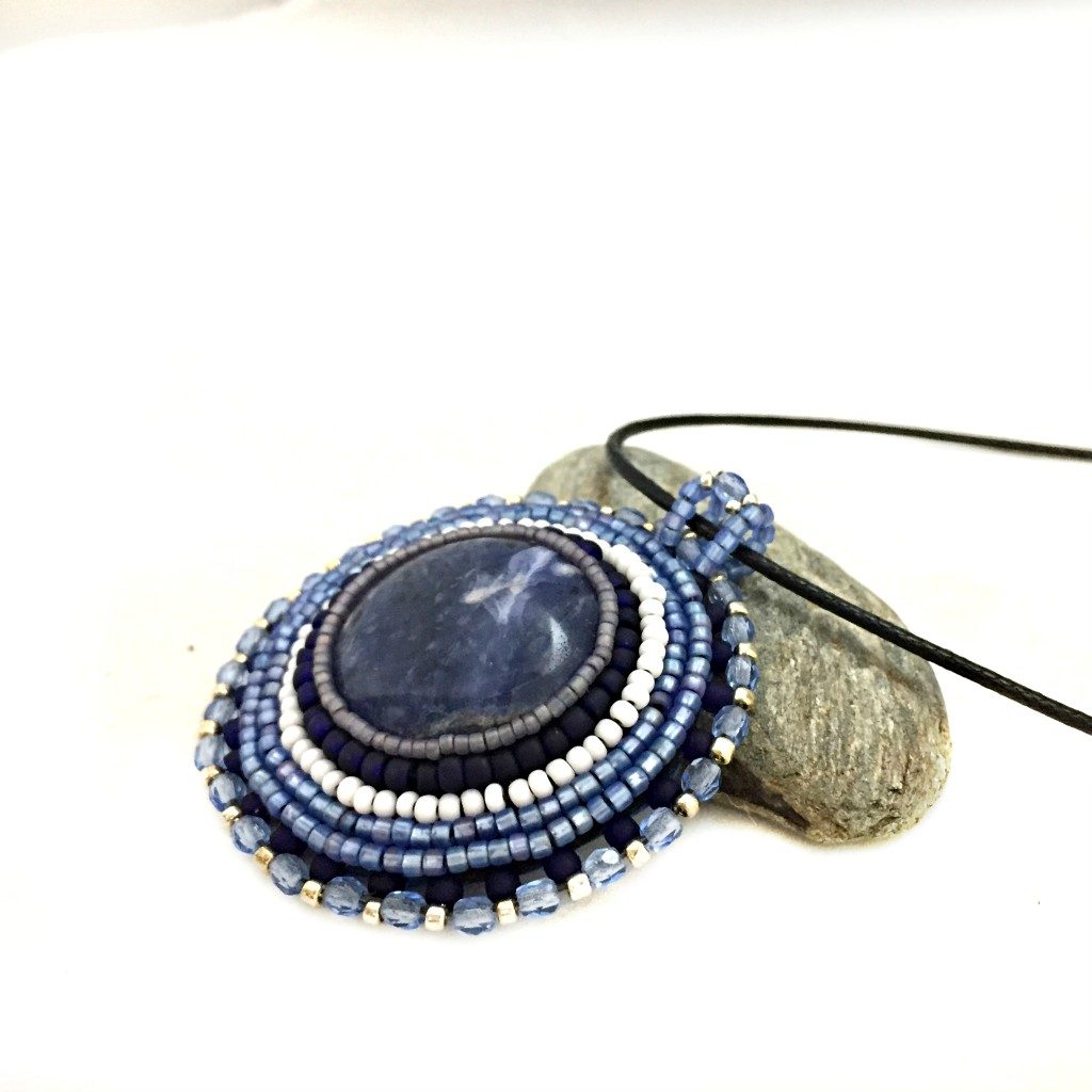 Side profile of round blue sodalite gemstone necklace with blue, silver and white seed bead edging. necklace is resting on a pebble.
