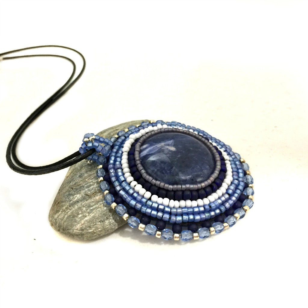 Side profile of round blue sodalite gemstone necklace with blue, silver and white seed bead edging. necklace is resting on a pebble.