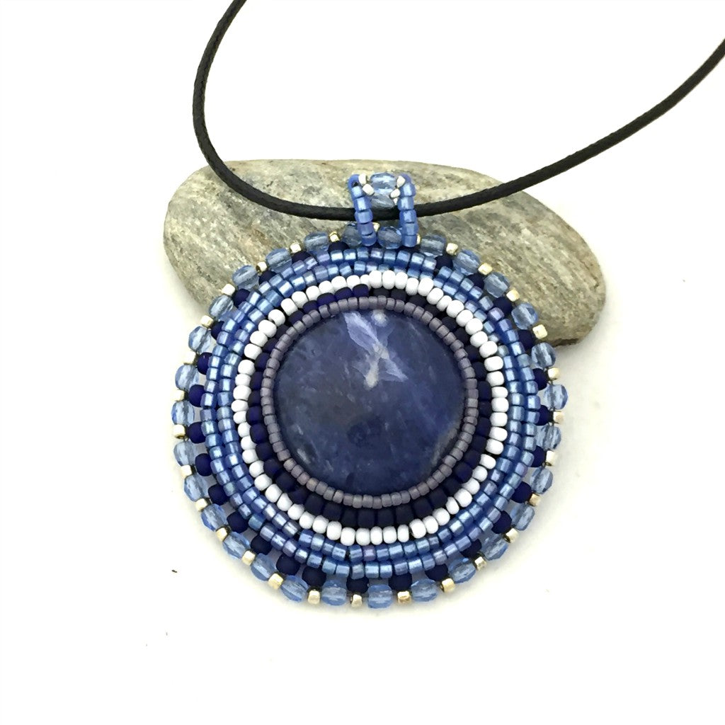 close up of round blue sodalite gemstone necklace with blue, silver and white seed bead edging. necklace is resting on a pebble.