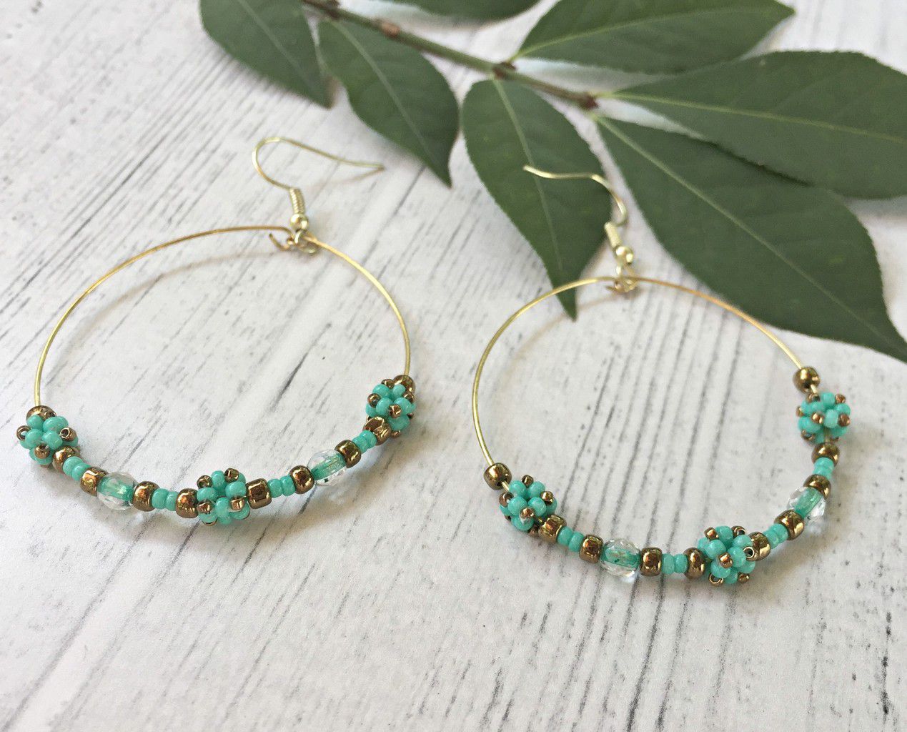 2 inch gold wire hoop earrings with turquoise cube beads, crystals, and seed beads laying on white washed wood with a leaf in the background.