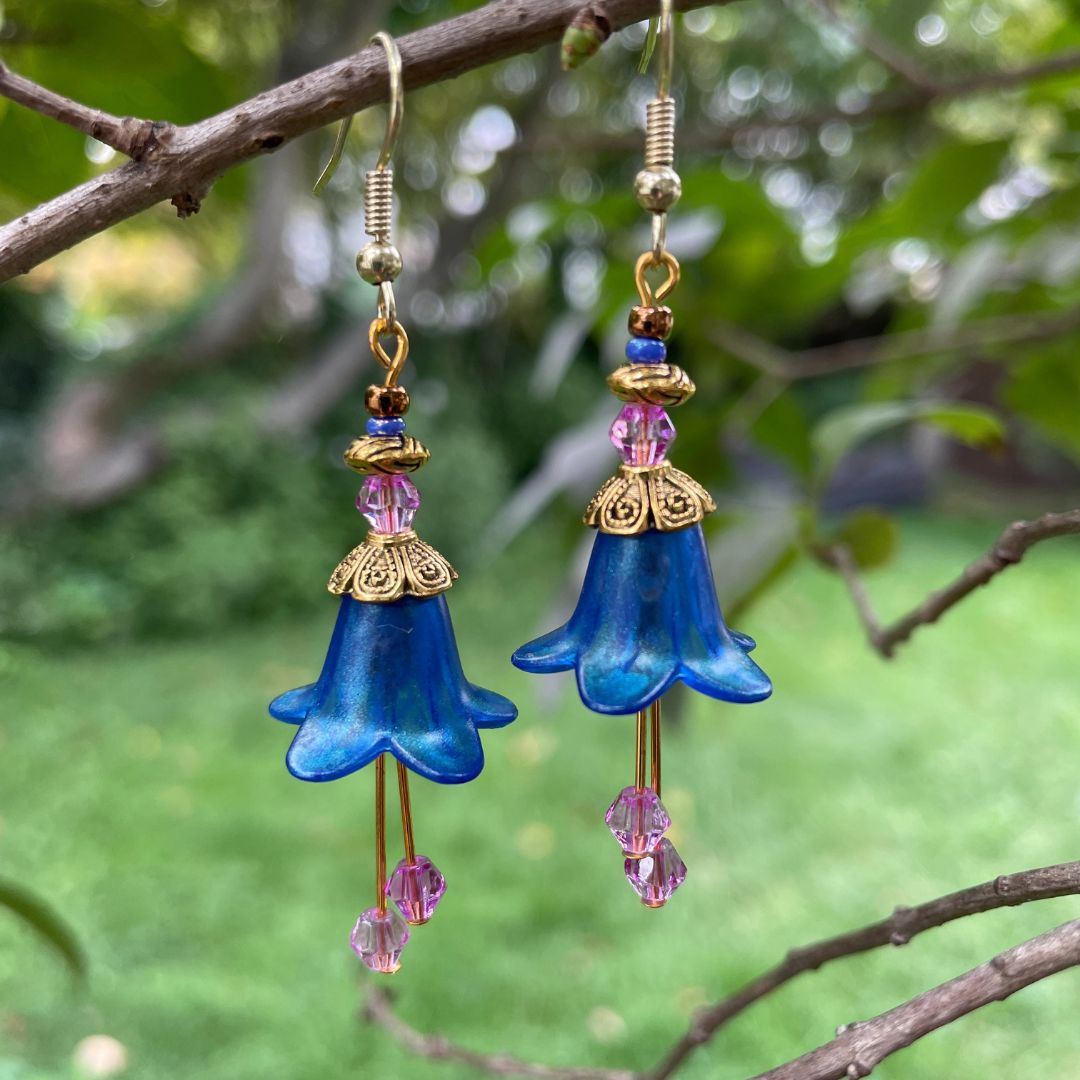 Blue flower dangle earring with gold accents and purple beads on a branch  in a garden.