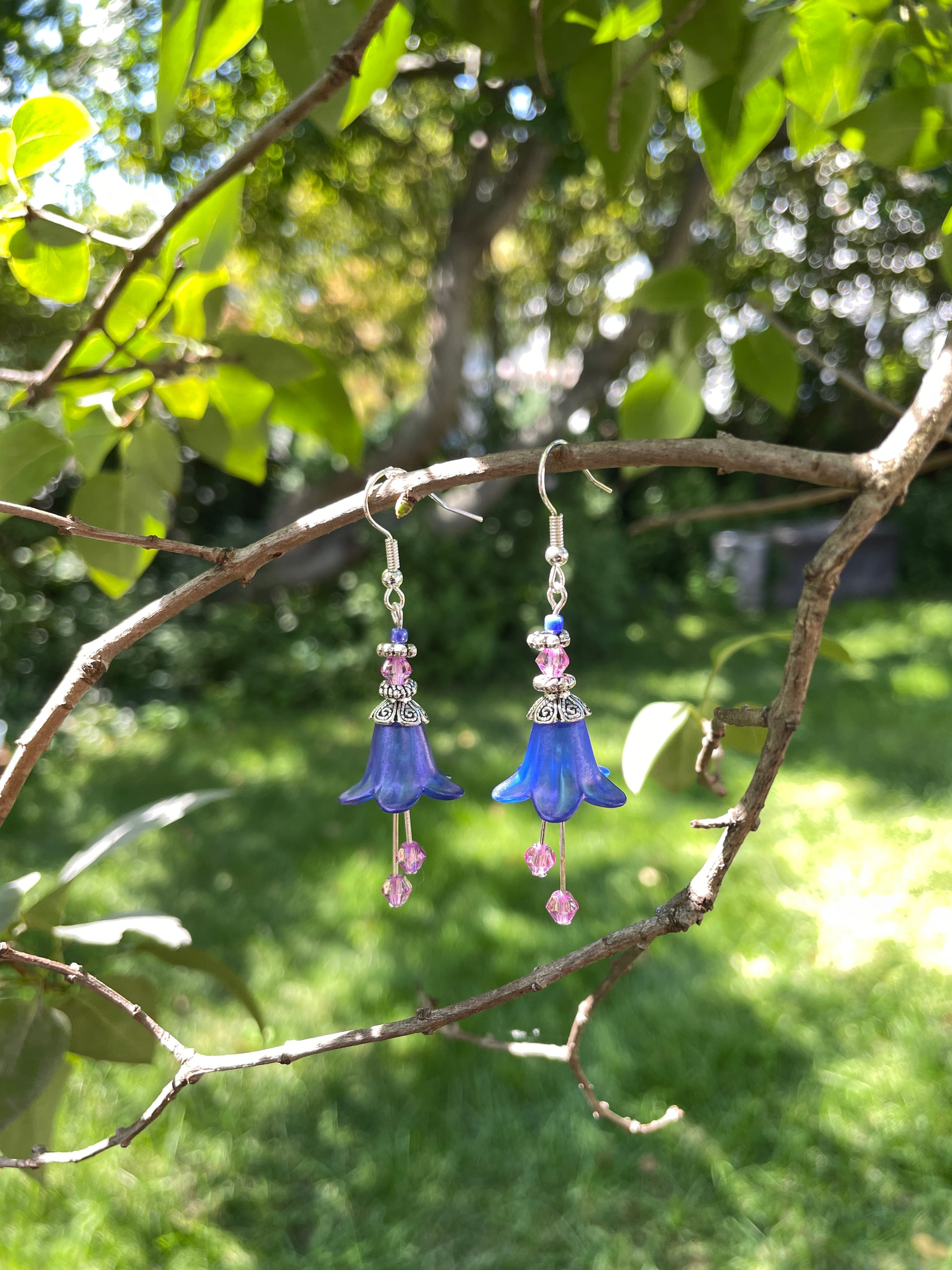 Blue flower dangle earring with silver and purple beads on a branch  in a garden.