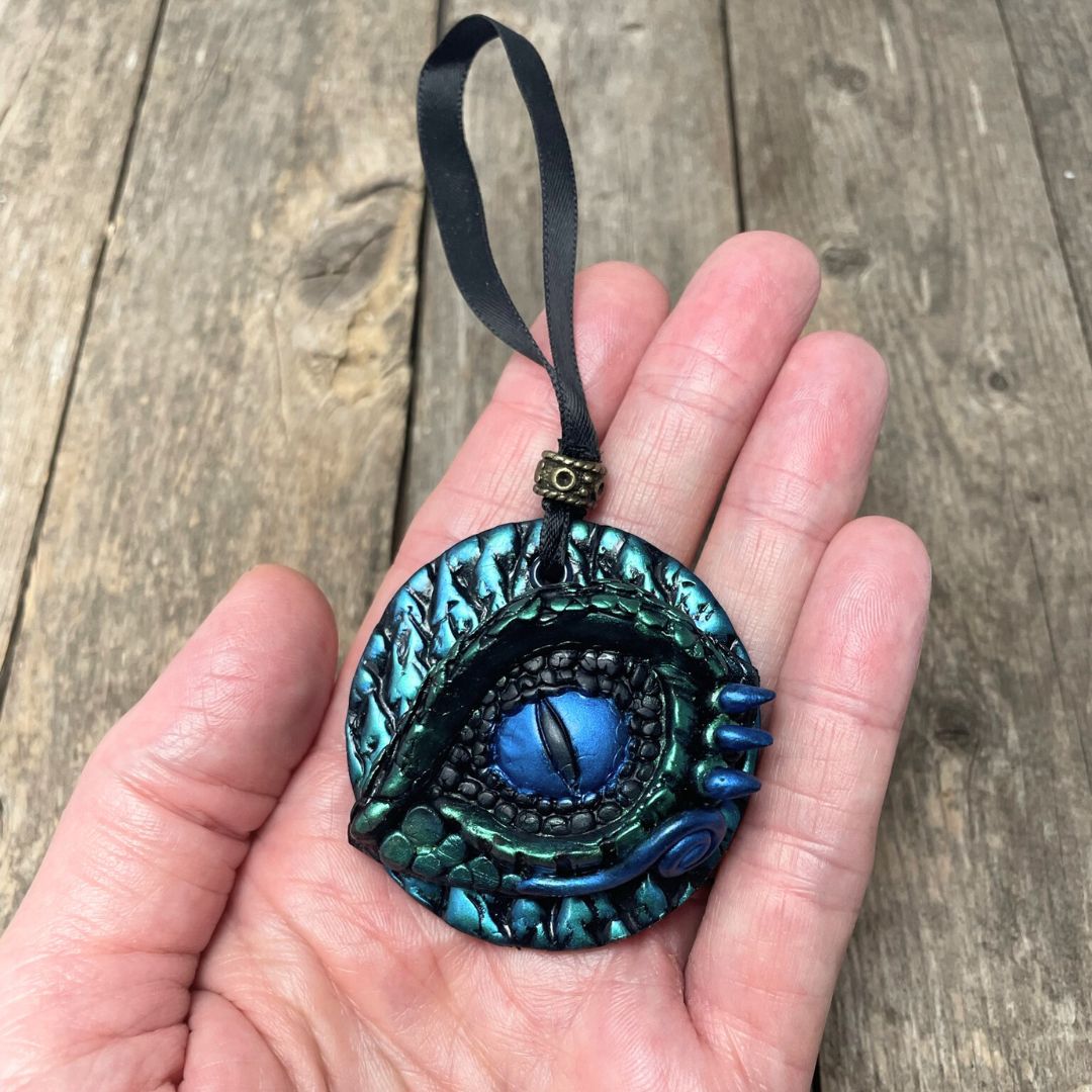 A round dragon eye ornament with metallic green and blue coloring with black satin ribbon held in a hand.