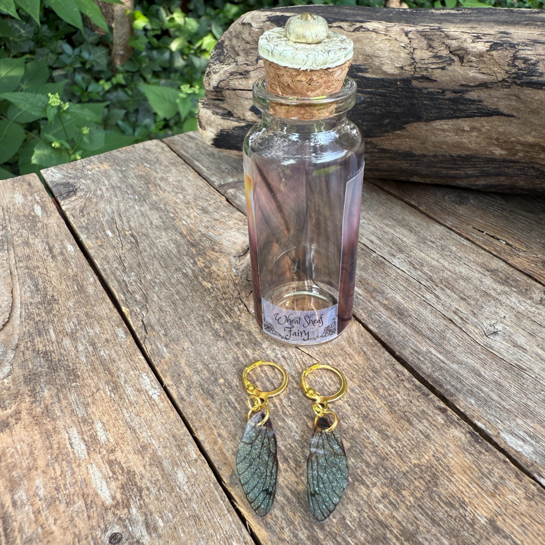 small fairy wing earrings lay flat on wood with small glass bottle .