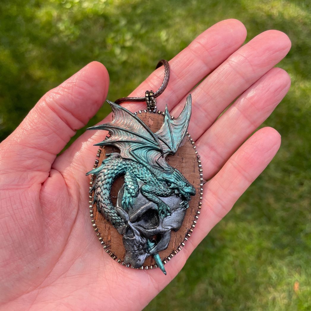 Oval pendant with a polymer clay green dragon upon a silver fierce skull. in hand.