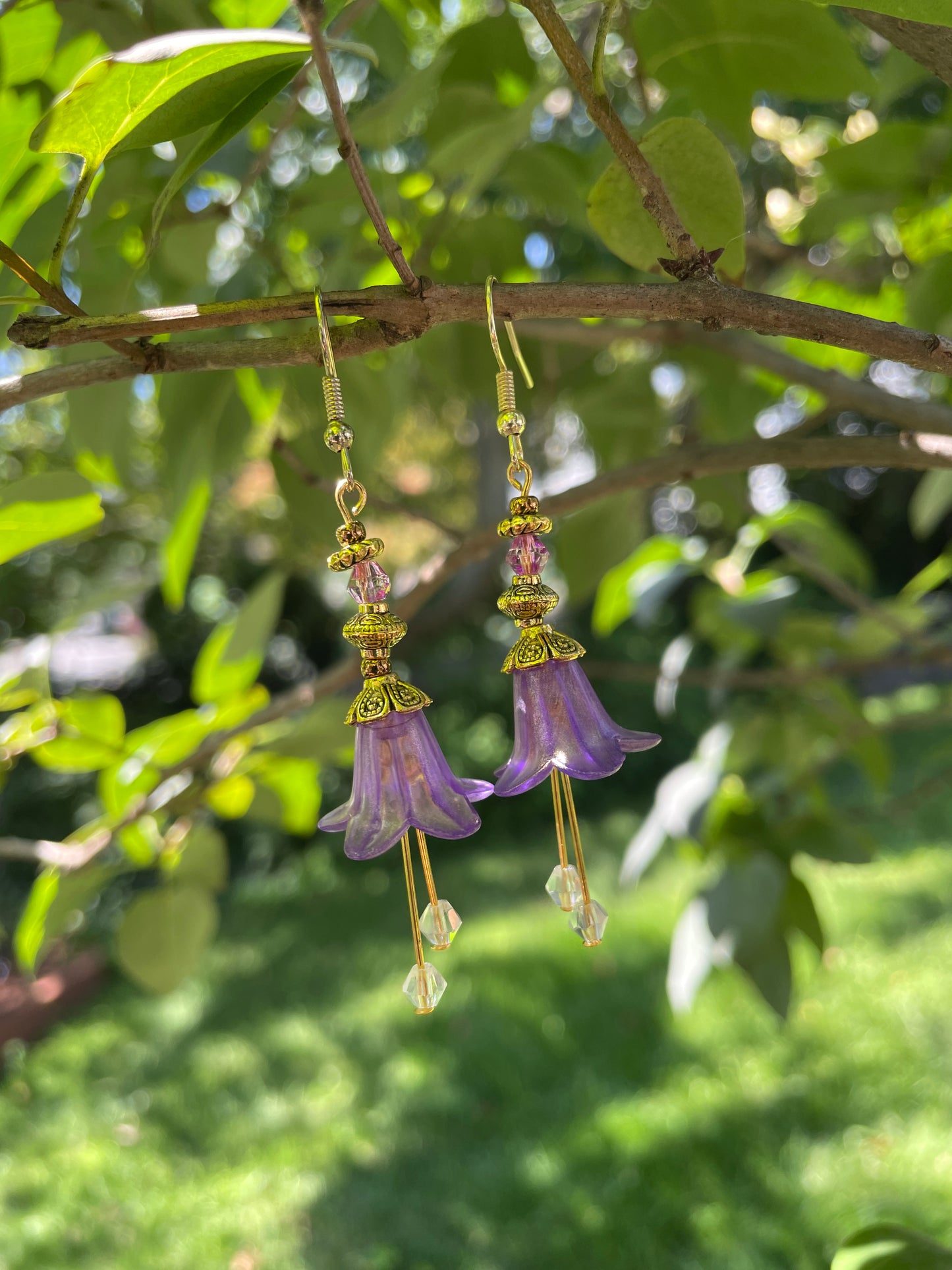 purple bell flower dangle earrings with clear crystal stamen and gold accent beads on a branch in a garden setting.