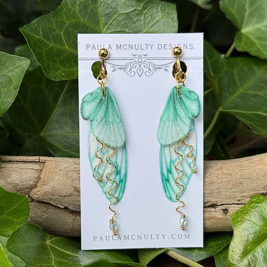 Close-up of mint green fairy wing earrings in gold finish showing glittered double wings and crystal accents.