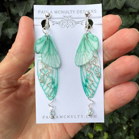 Fairycore mint green butterfly clip-on earrings resting on floral backdrop, perfect for fantasy jewelry lovers.