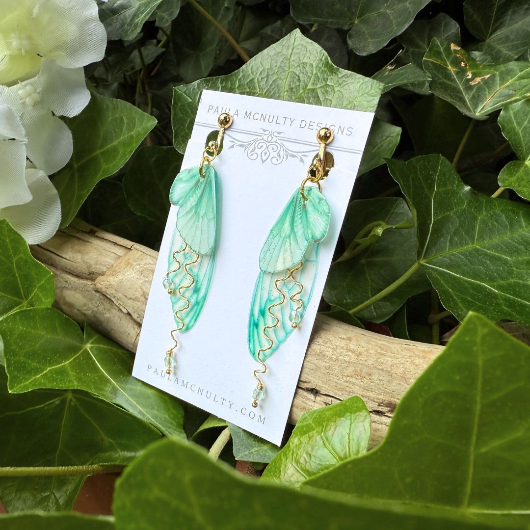 Close-up of mint green fairy wing earrings in gold finish showing glittered double wings and crystal accents.