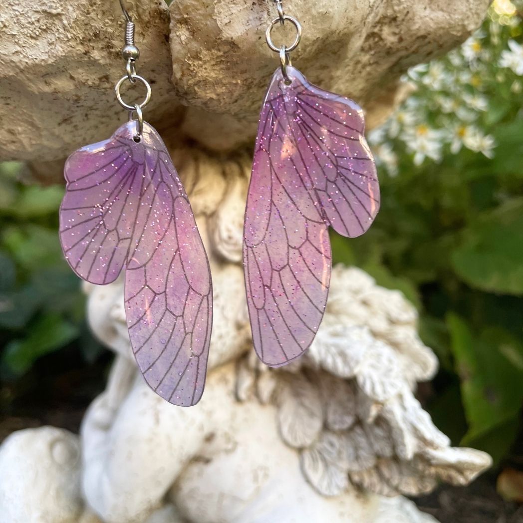 pink and purple sparkly glitter plastic fairy wing earrings hanging from a fairy garden ornament